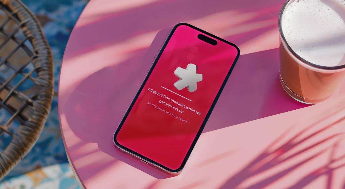 Smartphone with a pink screen displaying the Uptime app setup screen, placed on a pink round table with a glass of beer and a wicker basket in the background