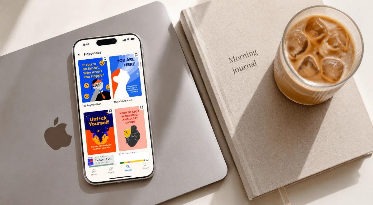 iPhone displaying a happiness app on a MacBook beside a morning journal notebook and iced coffee, representing a productive morning routine setup