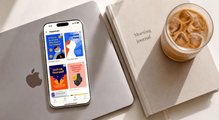 iPhone displaying a happiness app on a MacBook beside a morning journal notebook and iced coffee, representing a productive morning routine setup