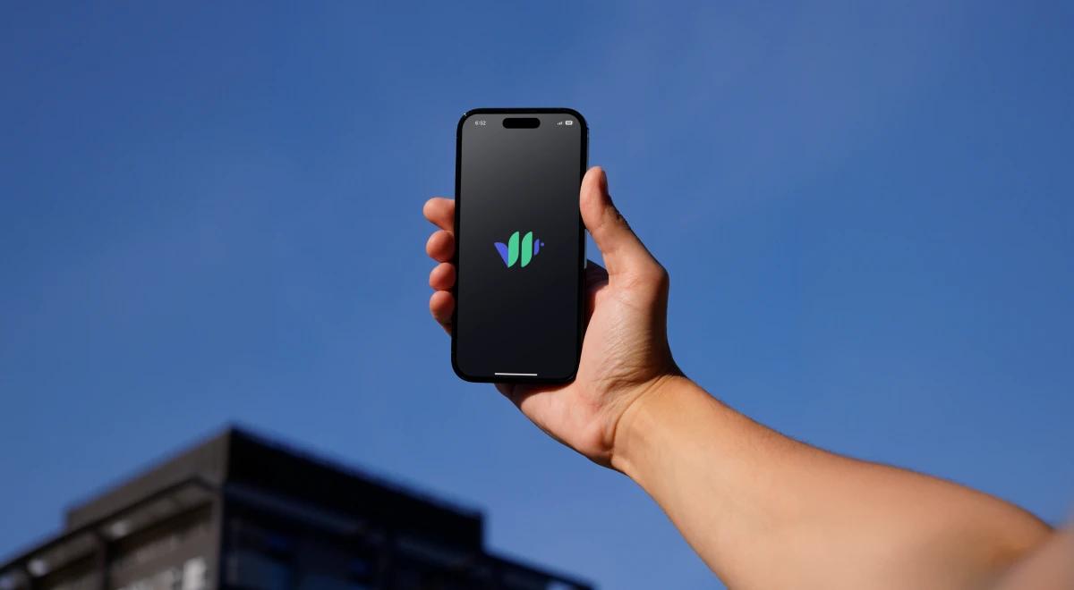 _Hand holding a smartphone with the Vocal Image app logo against a blue sky and building corner in the background