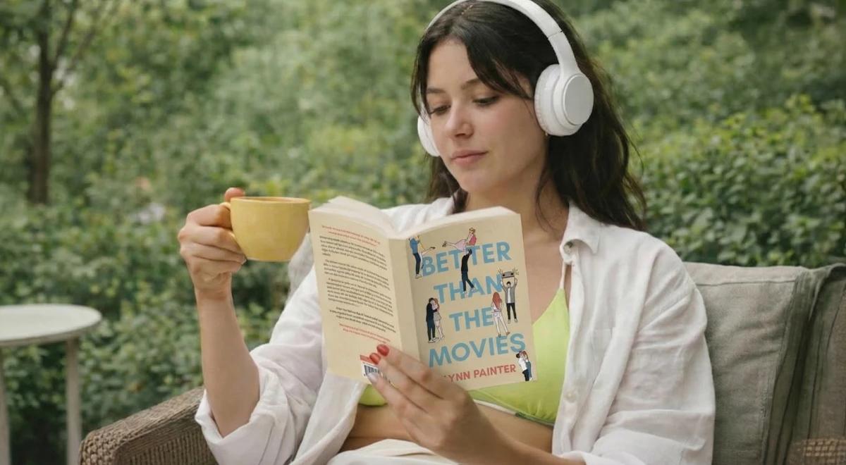 Woman with white headphones reading Better Than the Movies by Lynn Painter while holding a yellow cup outdoors among green trees, ideal for a rom-com audiobook review