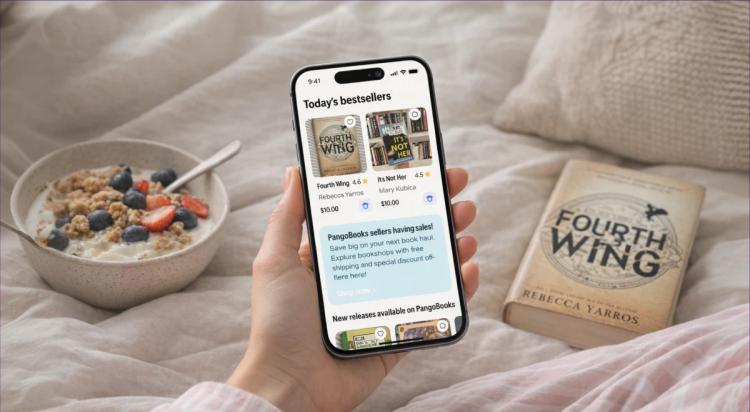 Hand holding a smartphone with the PangoBooks book app showing today's bestsellers, beside a bowl of granola and a physical copy of Fourth Wing by Rebecca Yarros on a cozy bed