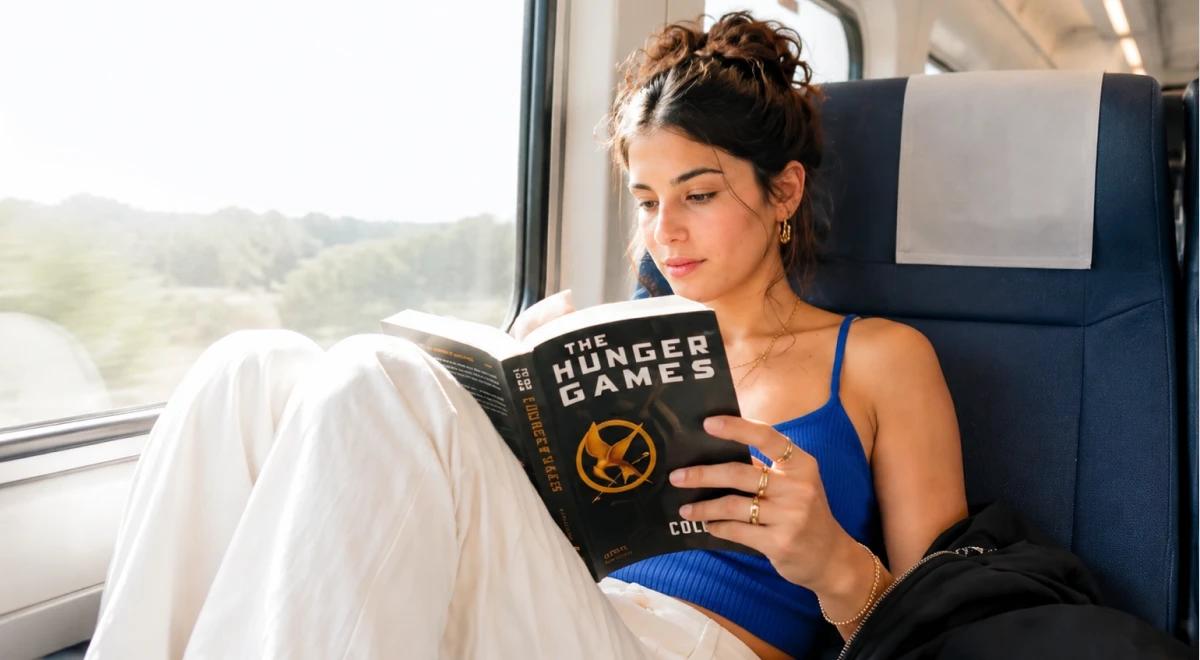 Young woman in a blue top reading The Hunger Games book while sitting in a train seat, with a green landscape visible through the window