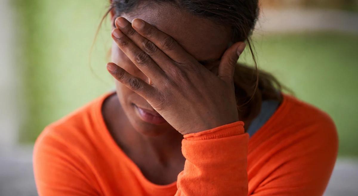 Woman in an orange shirt covering her face with her hands against a green blurred background, expressing emotional distress linked to anxious attachment style