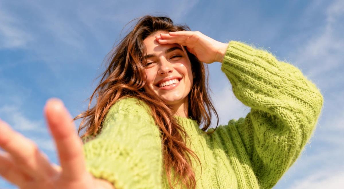 Smiling young woman in a lime green knit sweater shielding her eyes from the sun against a bright blue sky