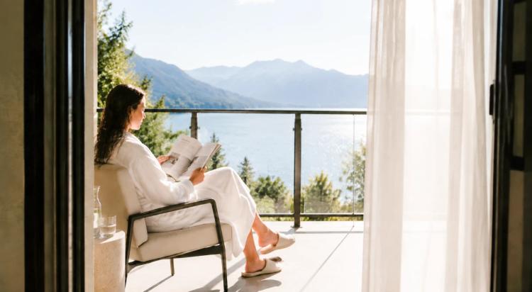 Woman sitting on a sunlit balcony reading, with a mountain lake view and sheer curtains in the background