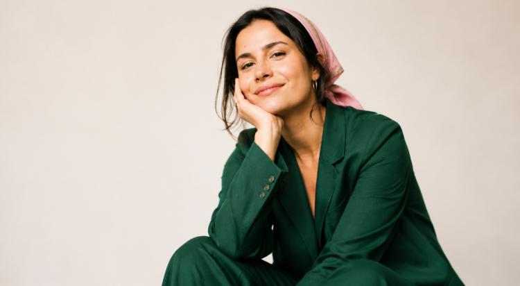 Smiling woman in a green blazer and pink headscarf resting her chin on her hand against a neutral beige backdrop, embodying be the woman he can't forget confidence