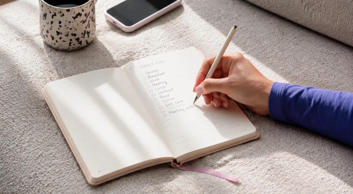 Hand writing a list in an open notebook next to a smartphone and speckled mug on a couch, representing discipline and goal-setting habits for motivation