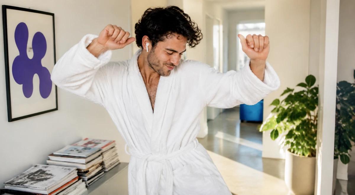 Happy man in a white bathrobe dancing freely in a bright living room with houseplants and art on the wall, expressing confidence and high self-esteem