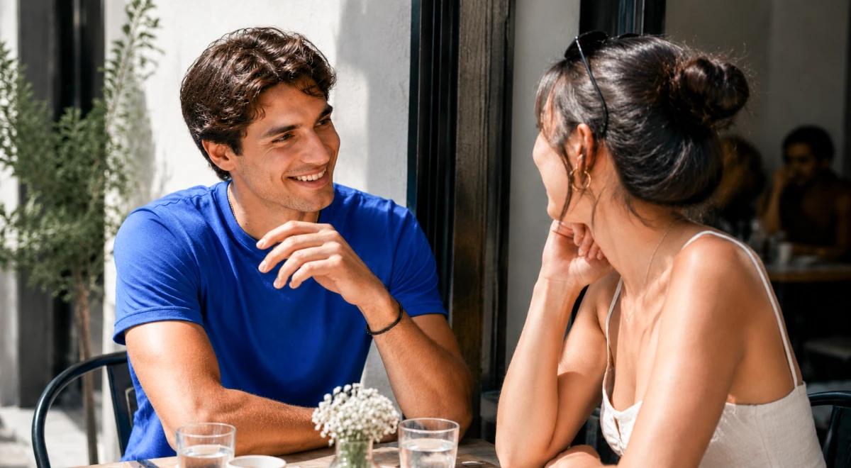 Young man in a blue t-shirt smiling and chatting with a dark-haired woman at an outdoor café table, illustrating a flirty in-person conversation after texting