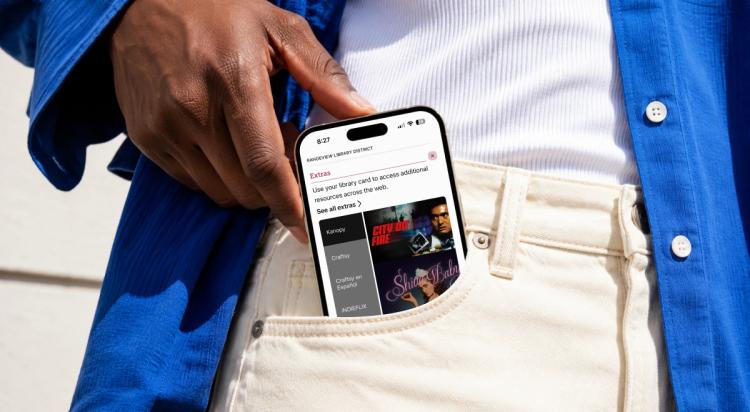 Person in a blue shirt pulling a smartphone with the Libby app open out of their pocket, showing digital library book covers outdoors