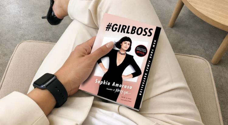 Woman wearing a black Apple Watch holding the #GIRLBOSS book by Sophia Amoruso while sitting on a beige couch, black heels visible in the background β self-growth read for a woman CEO