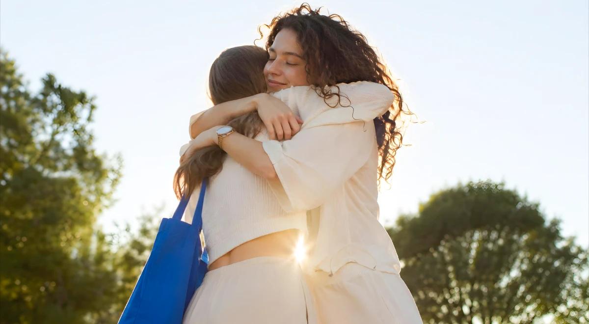 Two women sharing a warm embrace outdoors in sunlight with green trees in the background, representing forgiveness and emotional reconciliation