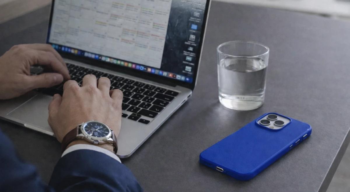 Entrepreneur in suit typing on MacBook with a busy calendar on screen, blue iPhone and glass of water on dark desk, illustrating Elon Musk daily routine and entrepreneurs time management