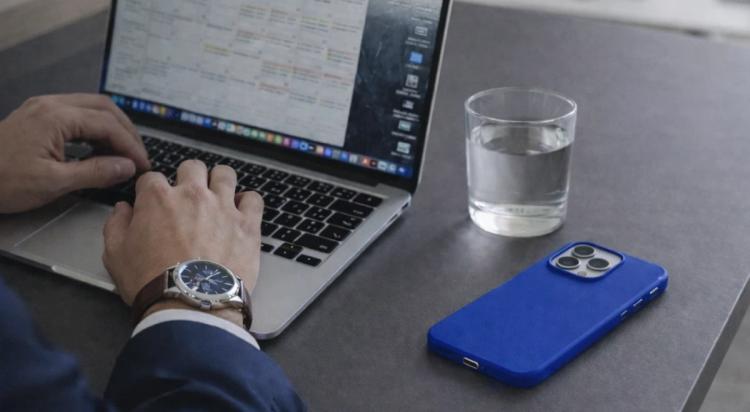 Entrepreneur in suit typing on MacBook with a busy calendar on screen, blue iPhone and glass of water on dark desk, illustrating Elon Musk daily routine and entrepreneurs time management