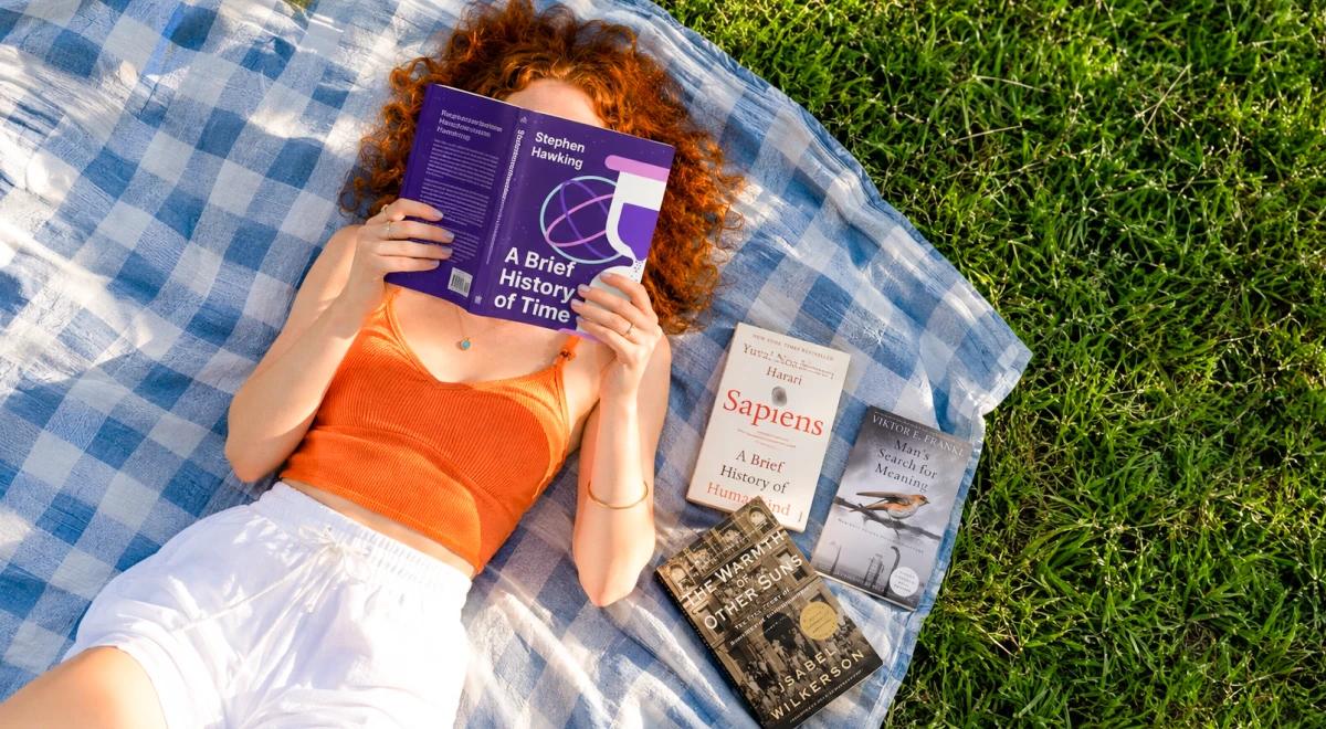 Young woman with curly red hair lying on a blanket on grass reading A Brief History of Time, with Sapiens and other nonfiction books beside her