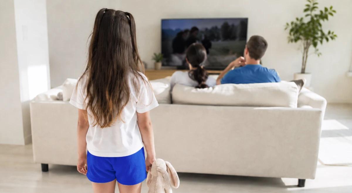 Young girl holding a stuffed bunny standing alone in a living room while her parents sit on a couch watching TV, illustrating child neglect and generational trauma