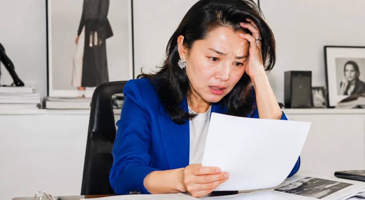 Stressed woman in a blue blazer sitting at a desk holding a document with a worried expression, hand on her forehead, with framed photos and fashion artwork on the wall behind her