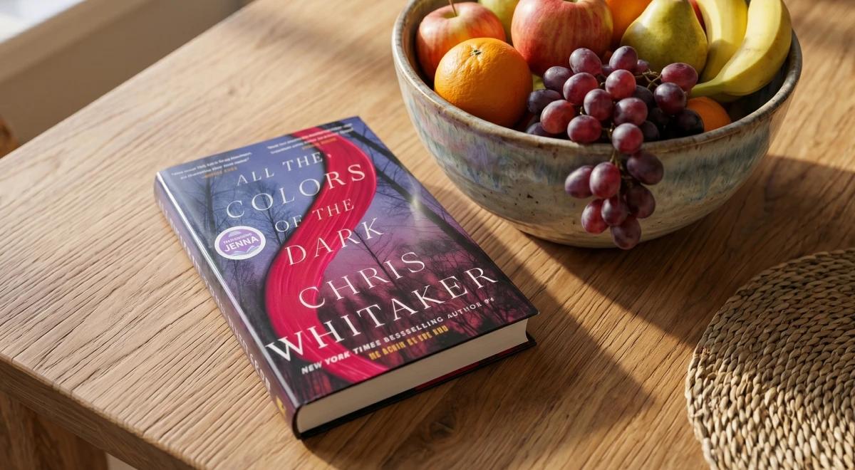 All the Colors of the Dark by Chris Whitaker paperback on a wooden table beside a bowl of mixed fruit, representing a popular fiction book summary