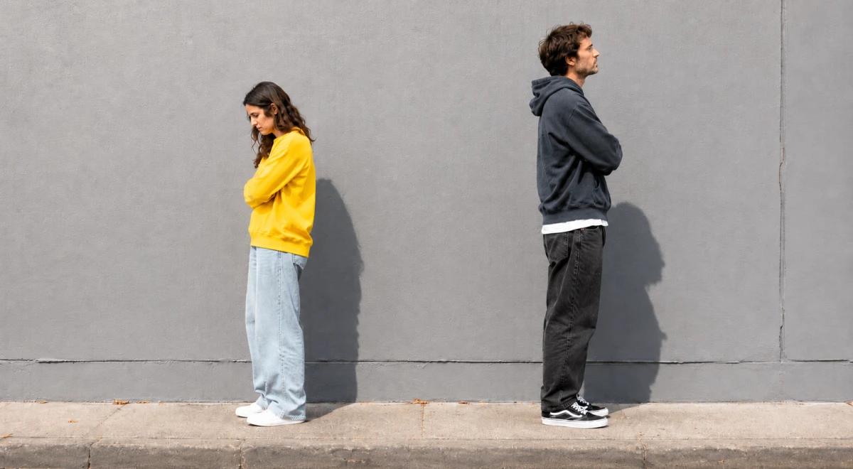 _A woman in a yellow hoodie and a man in a dark hoodie standing back to back against a gray wall, representing conflict and emotional distance in a relationship