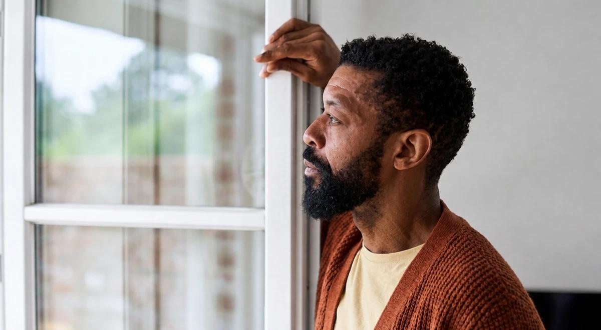 Thoughtful Black man in a rust orange cardigan leaning against a window frame looking outside, reflecting on existential thoughts in a dimly lit room