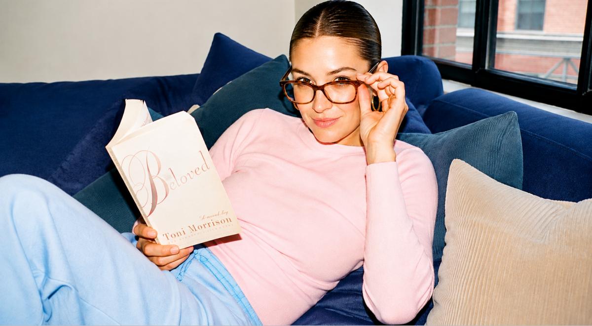 Woman in a pink top and glasses relaxing on a blue velvet sofa while holding Beloved by Toni Morrison, embodying a hot person reading list by a sunlit city window