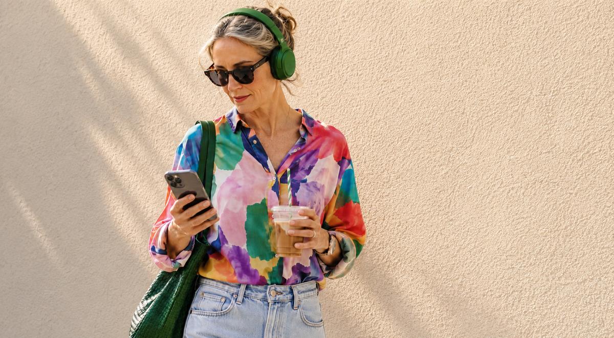 Woman in green headphones and a colorful blouse holding an iced coffee and smartphone against a beige wall, ready to listen to 20 books in 20 days
