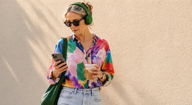 Woman in green headphones and a colorful blouse holding an iced coffee and smartphone against a beige wall, ready to listen to 20 books in 20 days