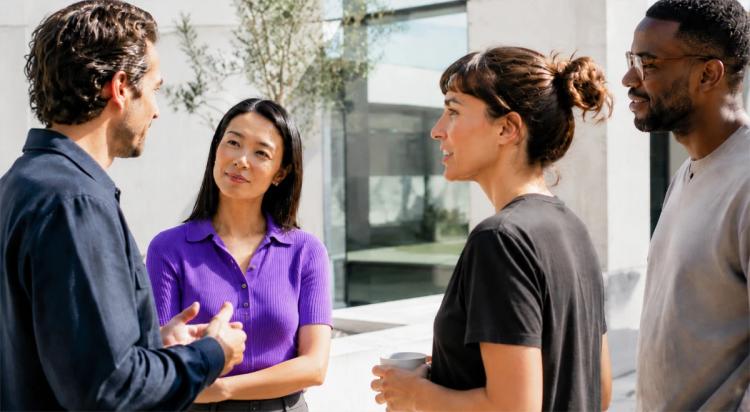 Four colleagues engaged in an outdoor conversation in small groups, with a woman in a purple top actively speaking, showing strong interpersonal communication and high EQ skills