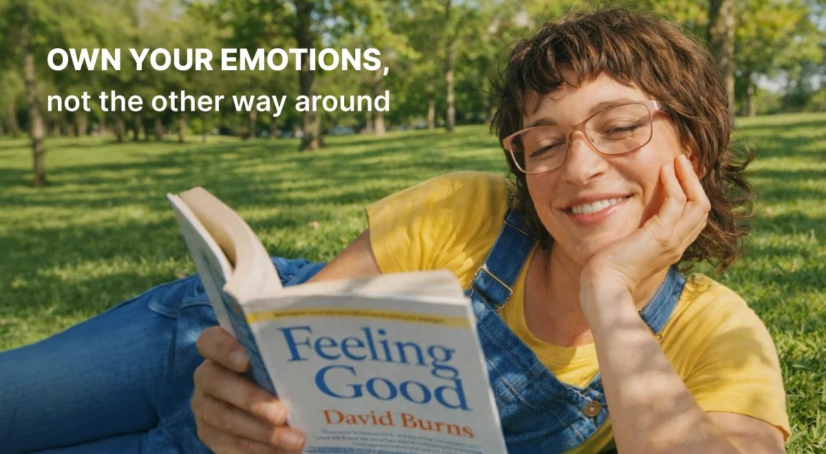 Smiling woman in yellow shirt and denim overalls lying on green grass reading Feeling Good by David Burns, a top book recommendation for managing emotions outdoors