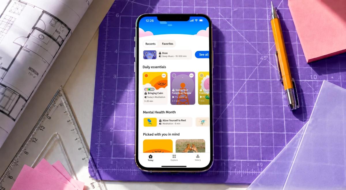 iPhone displaying a wellness app home screen with daily essentials and mental health content, placed on a purple cutting mat surrounded by blueprints, ruler, pencil, and pink paper