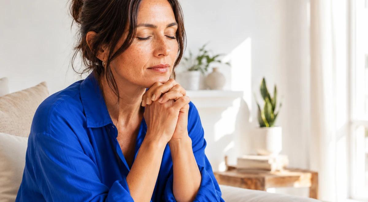 Woman in a blue shirt sitting with closed eyes and hands clasped in a bright bedroom with plants, practicing mindfulness and inner calm