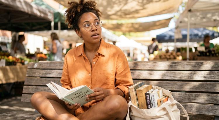 Woman with coily hair and glasses reading _The Financial Diet_ paperback cross-legged at sunny outdoor market, canvas tote of best books for beginners beside her, warm golden light 1