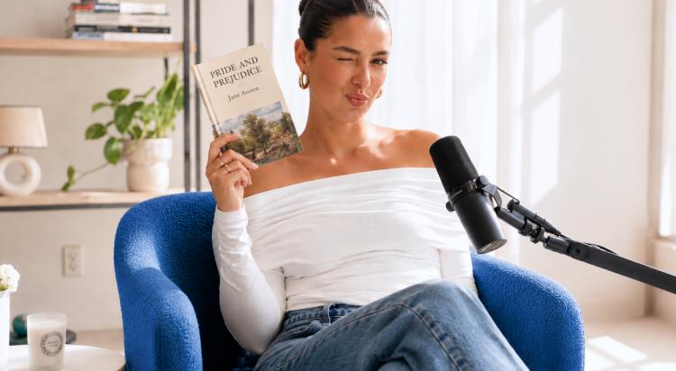 Woman winking while holding Pride and Prejudice by Jane Austen during a book trivia podcast, seated in a blue armchair beside a microphone in a bright room with plants and shelves