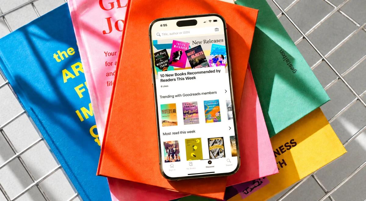 iPhone displaying a reading app screen resting on a pile of colorful books and tote bags on a grid surface