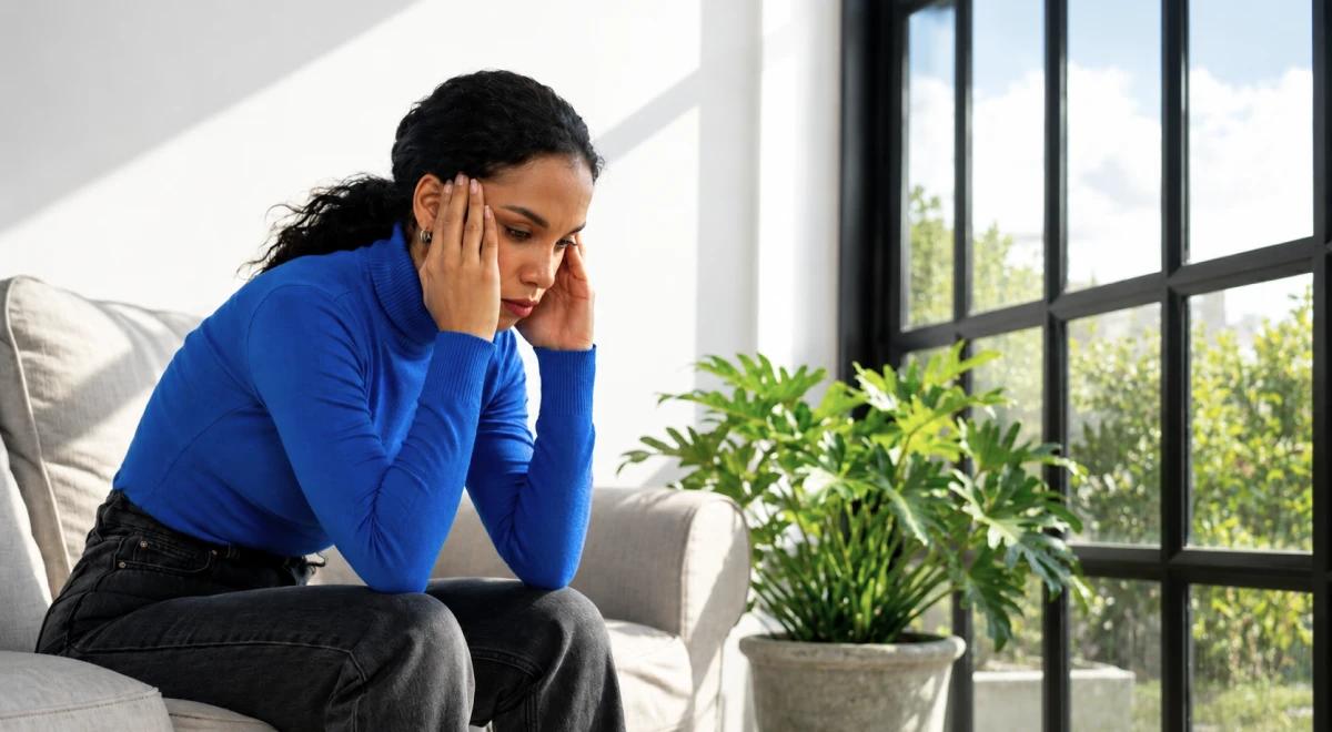 Young woman in a blue turtleneck sitting on a couch holding her head with a worried expression, appearing hyperaware and anxious in a bright living room with green plants and large windows