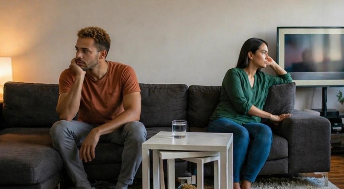 Couple sitting apart on a gray sofa in a dimly lit living room, avoiding eye contact, showing signs of relationship burnout and emotional distance