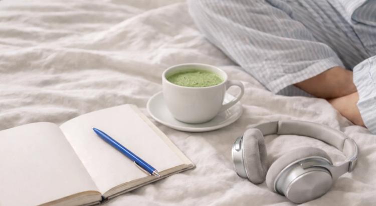 Person in bed with open notebook, blue pen, cup of green matcha, and silver headphones on white linen as part of a successful person's morning routine