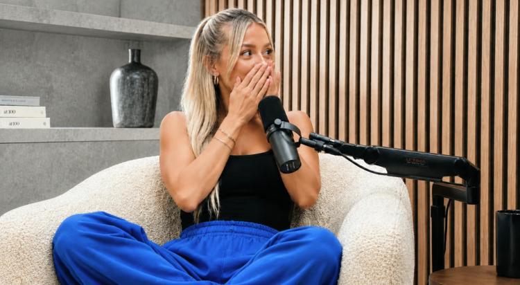 Blonde woman in black top and blue pants covering her mouth in surprise while sitting in a white chair next to a podcast microphone in a modern studio