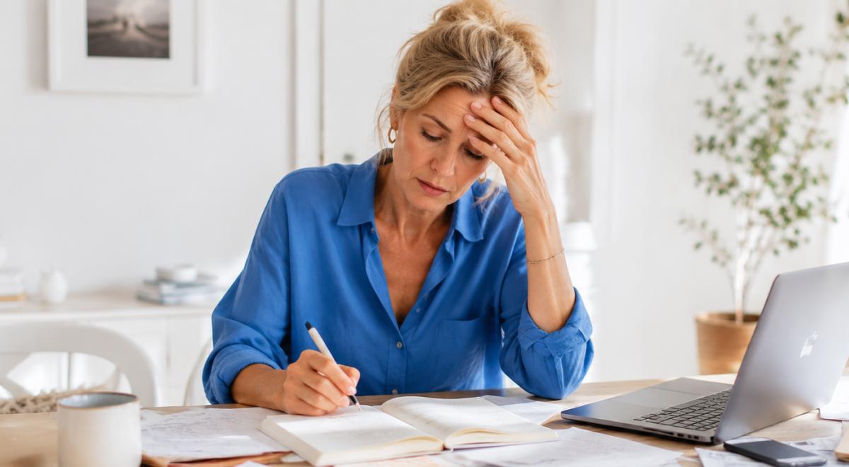 Stressed middle-aged woman in a blue shirt writing in a notebook at a home desk with a laptop, showing signs of adult ADHD focus struggles