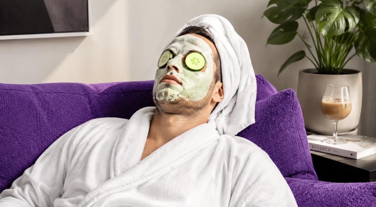 Man in white bathrobe and towel relaxing on a purple sofa with a clay face mask and cucumber slices on eyes, enjoying a self-care routine at home