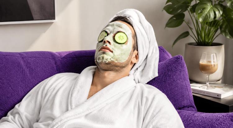 Man in white bathrobe and towel relaxing on a purple sofa with a clay face mask and cucumber slices on eyes, enjoying a self-care routine at home