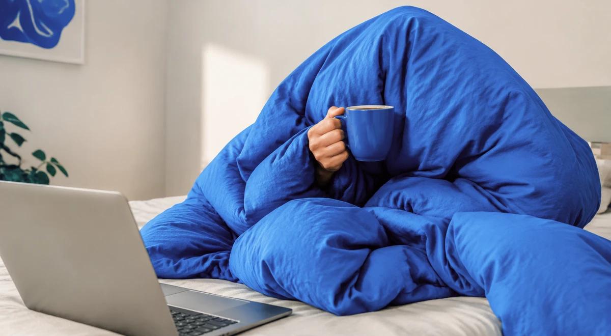 Person hiding under a blue blanket holding a mug while a laptop sits open on the bed, illustrating avoidance and self-sabotage behavior