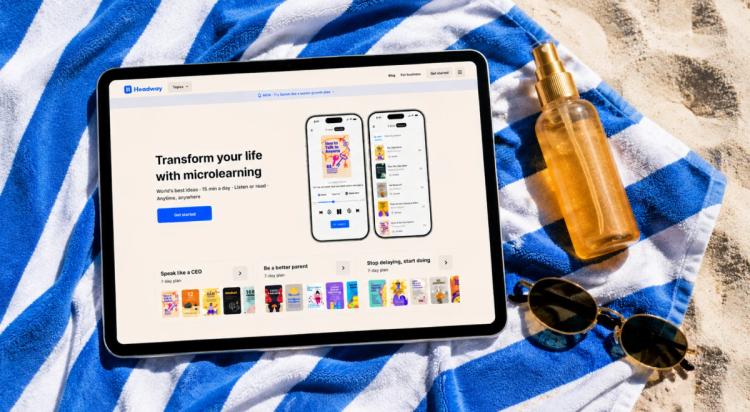 Headway app displayed on a tablet and smartphone lying on a blue and white striped beach towel with sunglasses and a sunscreen bottle on sandy background