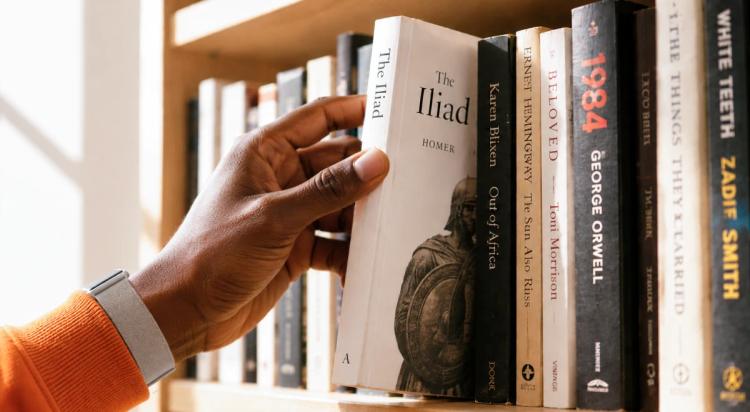 A hand in an orange sleeve pulling The Iliad by Homer from a wooden bookshelf filled with classic novels including 1984 by George Orwell and Beloved by Toni Morrison