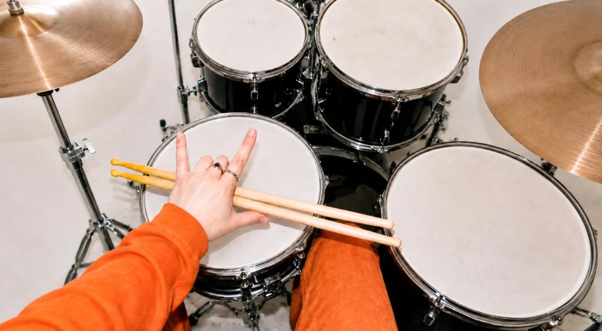 Drummer in an orange sleeve making a rock hand sign over a snare drum, holding wooden drumsticks, with a black drum kit and cymbals in the background