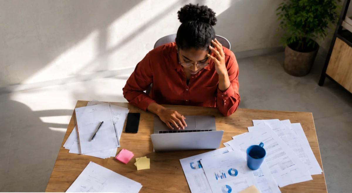 Stressed Black woman in a red shirt sitting alone at a desk covered with papers, charts, and sticky notes while working on a laptop in a bright office