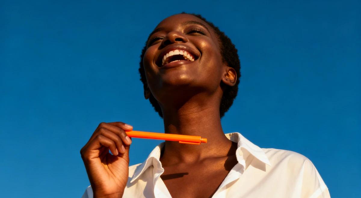 Confident smiling woman in a white shirt holding an orange pen against a bright blue sky, symbolizing CEO ambition and leadership mindset