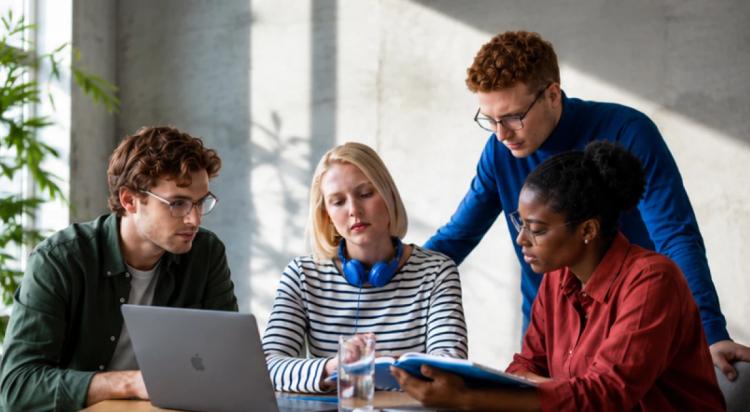 _Four diverse colleagues collaborating around a MacBook and tablet at an office table, engaged in a teamwork discussion with plants in the background