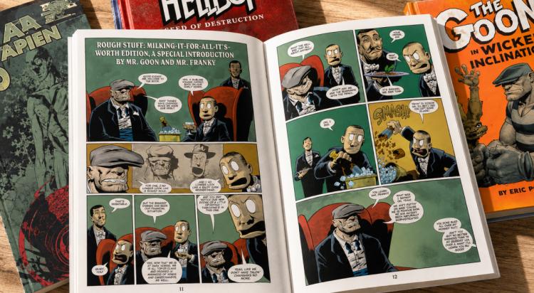 Open comic book showing colorful illustrated panels of characters in suits, surrounded by hardcover graphic novels like Hellboy and The Goon, ideal for comic book trivia fans